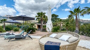 Un fauteuil de jardin en osier avec des coussins, un parasol et une chaise longue avec une petite table, le tout installé sur une terrasse agrémentée de palmiers et offrant une vue sur les montagnes.