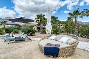 A wicker lounge chair with cushions, a covered umbrella, and a sun lounger with a table, all on a patio with palm trees and a mountain backdrop.