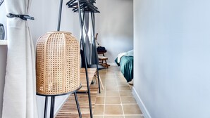 Un couloir étroit avec une chaise en rotin, un étendoir métallique et un lit recouvert d’une couverture verte.