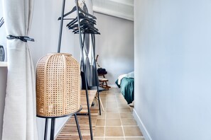 Un pasillo estrecho con una silla de ratán, un tendedero de metal y una cama con una colcha verde.