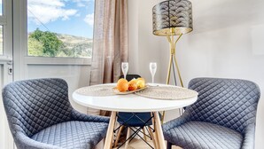 Une pièce avec deux fauteuils gris capitonnés, une table ronde blanche avec un set de table, une lampe décorative et une vue sur l’extérieur par une fenêtre.