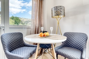 A room with two grey quilted chairs, a round white table with a placemat, a decorative lamp, and a view of the outdoors through a window.