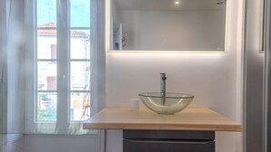 Une salle de bain avec un lavabo en verre, un miroir et une fenêtre donnant sur des immeubles.