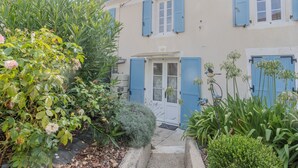 Un bâtiment avec des volets bleus, une porte et un petit jardin agrémenté de plantes et d’un pot de fleurs.