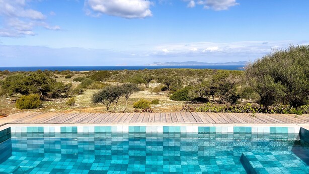 Une piscine aux carreaux bleu clair, entourée d’une terrasse en bois, offrant une vue sur un paysage arboré et, au loin, sur la mer.