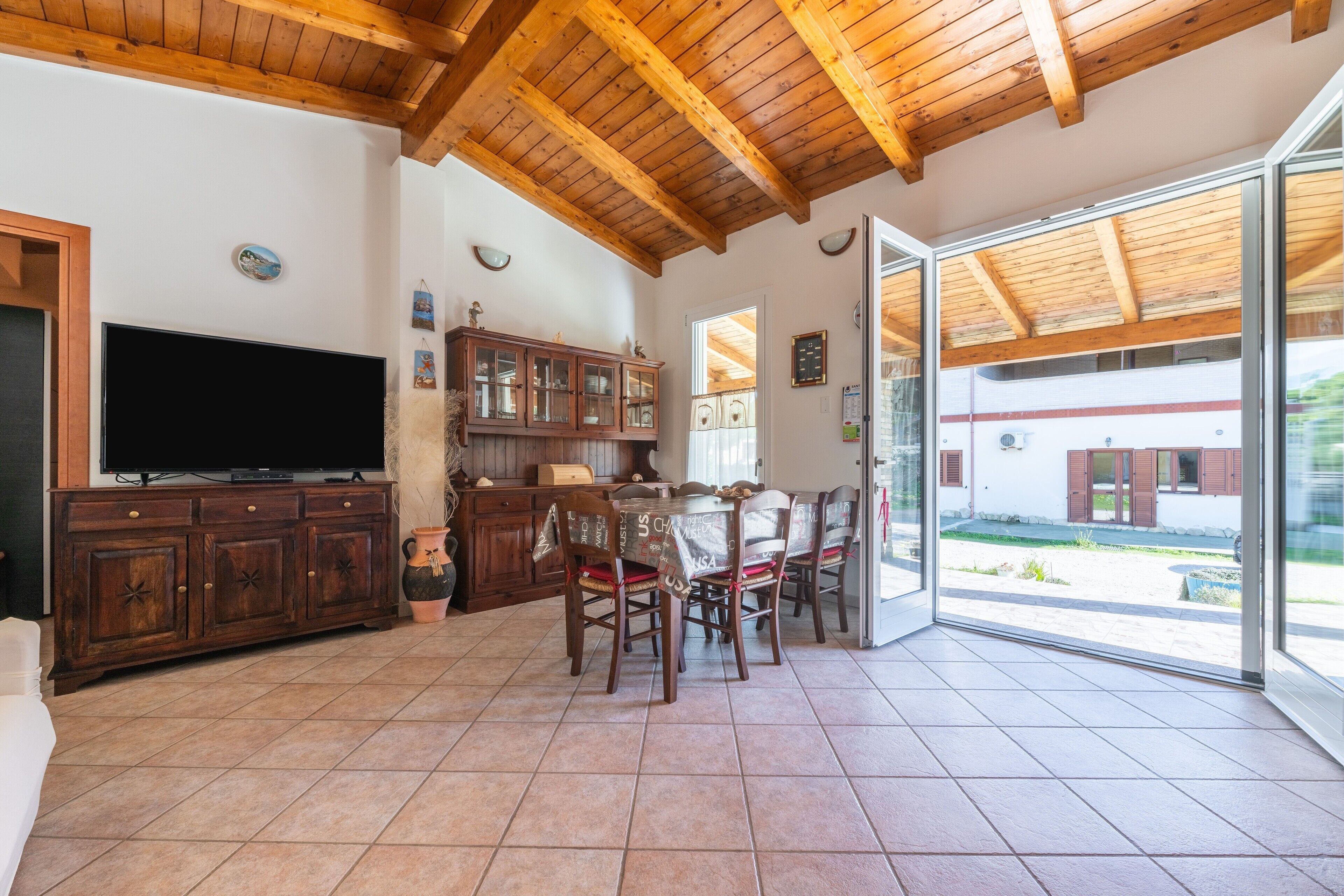 Amplia sala de estar con techo de madera, televisor, mesa de comedor con sillas y vistas a través de grandes puertas de vidrio.