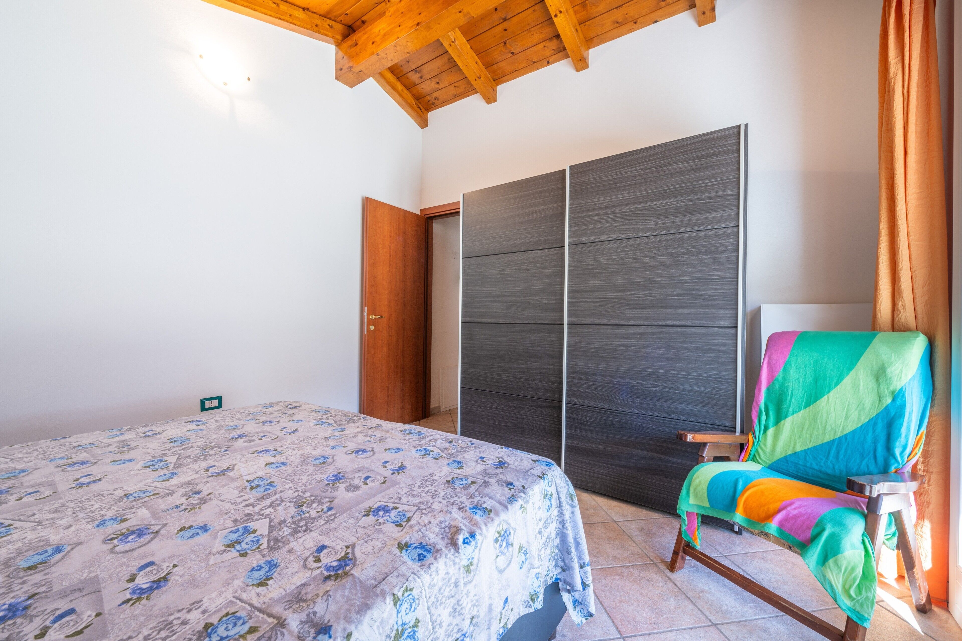 Un dormitorio con cama, armario, una silla colorida y techo de madera.