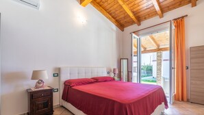 Een slaapkamer met een bed, nachtkastje, lamp, raam met gordijnen en uitzicht.