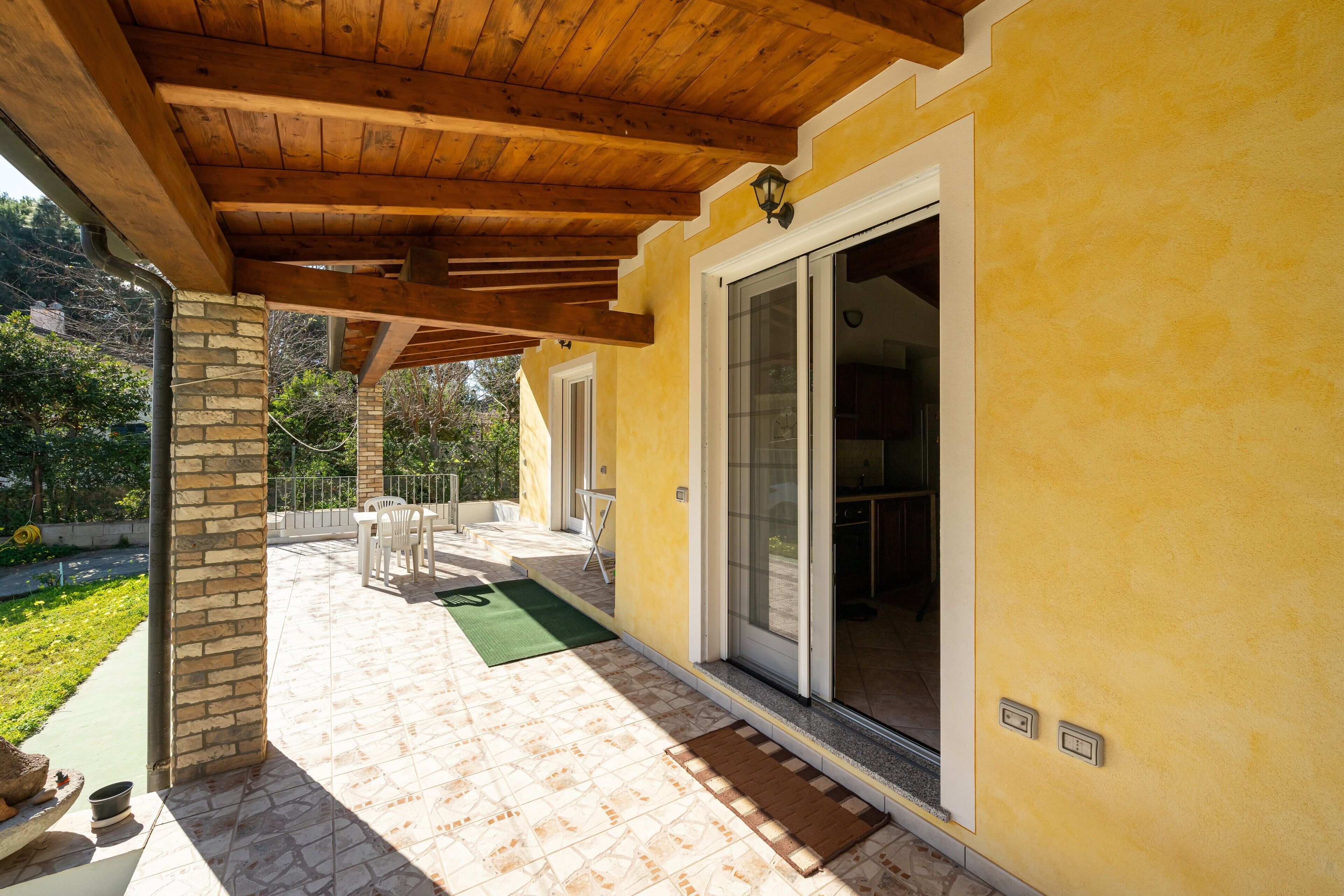 A covered patio with a stone column, a green mat, and a wooden table and chairs.
