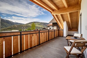 A wooden balcony with a view of mountains, a table with chairs, and a stone floor.