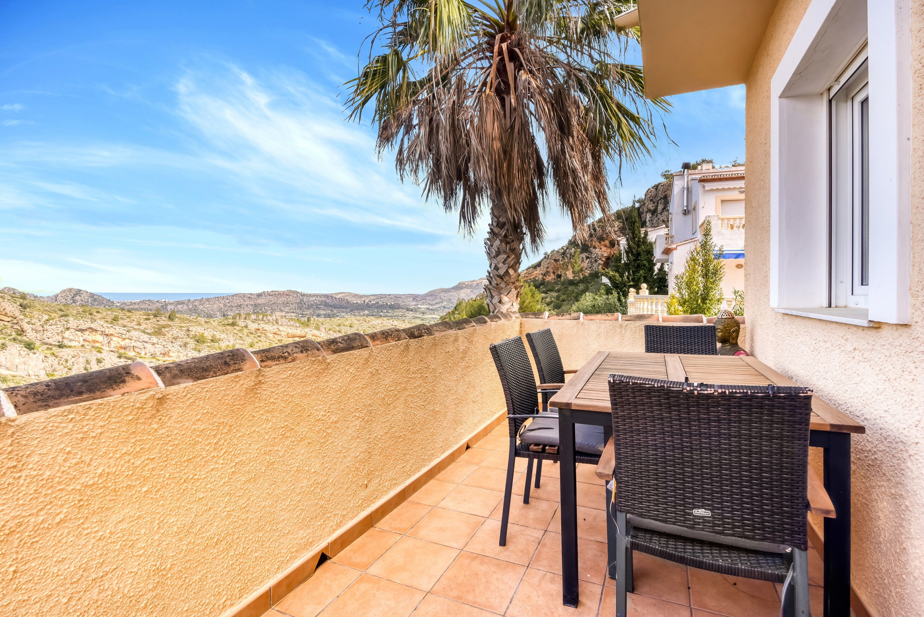 A terrace with a table and chairs, a palm tree, and a view of a hilly landscape.