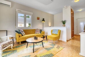 Een woonkamer met een geel sofa, een rond bijzettafeltje, een witte fauteuil, een klein tafeltje, een vloerlamp en een potplant.