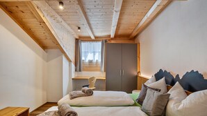 Ett sovrum med ett sluttande tak, en säng med kuddar och ett täcke, ett träbord som sängbord, en garderob och ett fönster med gardiner.