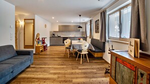 Una sala de estar moderna con sofá, mesa de comedor y sillas, una cocina al fondo, y un ventanal con cortinas.