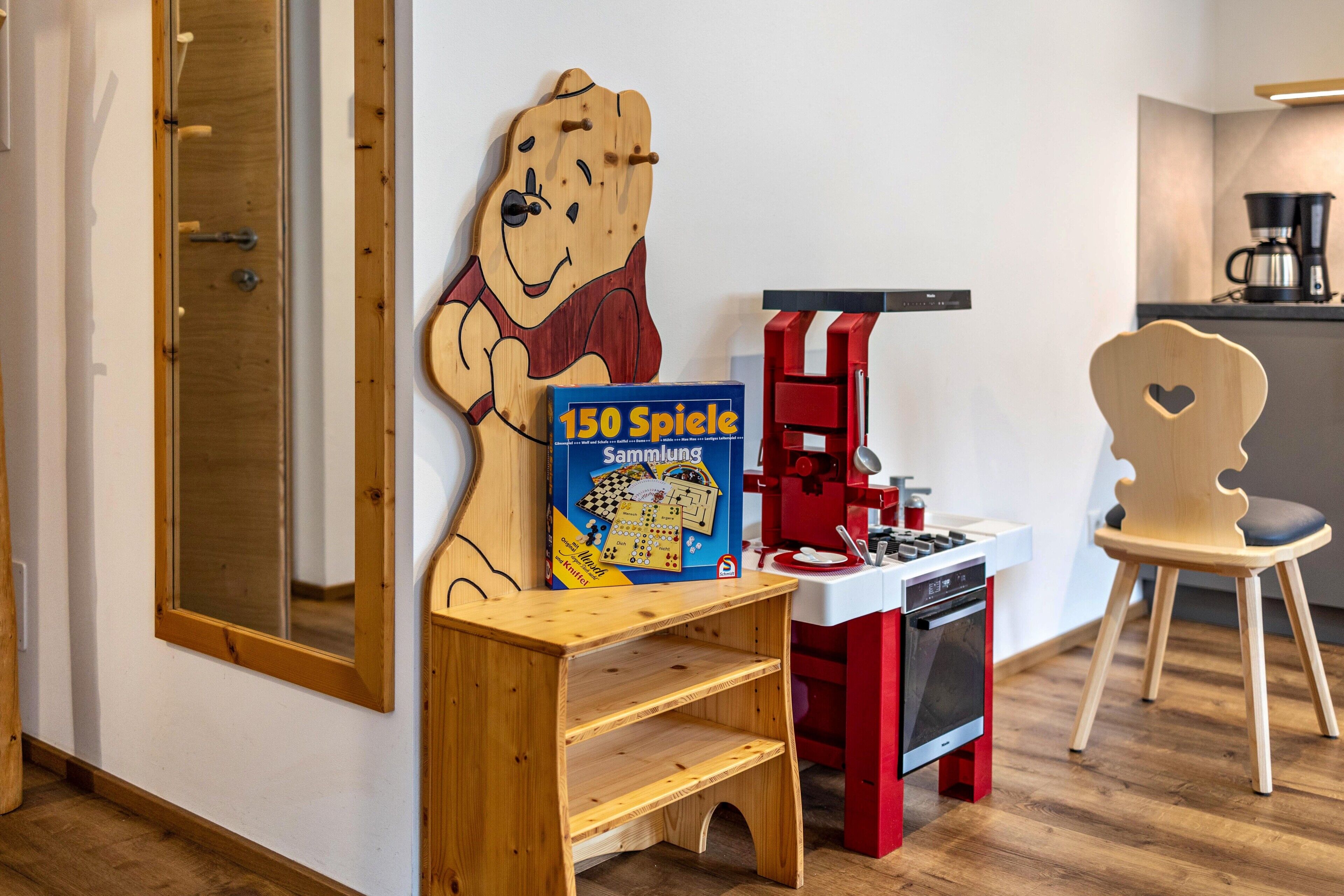 A children's play area with a wooden bear decoration, a table with a game box, a red toy kitchen, and a wooden chair.