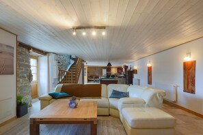 A spacious living area with a wooden ceiling, a stone wall, a staircase, a kitchen in the background, and a sofa with cushions.