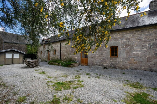 A stone building with a gravel courtyard, wooden furniture, and a tree with yellow flowers.