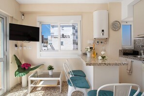 Une cuisine compacte dotée d’un four encastré, d’une fenêtre donnant sur des immeubles, d’un téléviseur à écran plat et d’un petit coin repas avec des chaises.