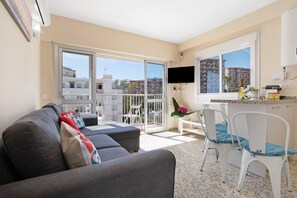 Un appartement moderne avec un canapé gris, des chaises blanches, un téléviseur et un balcon offrant une vue sur les immeubles.