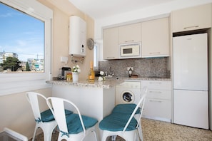 A compact kitchen with a washing machine, a microwave, and a refrigerator. There are two chairs with teal cushions.