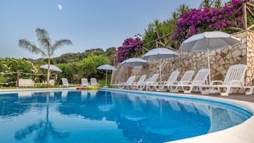 Ein Swimmingpool mit Liegestühlen und Sonnenschirmen, umgeben von Steinmauern und üppiger Vegetation.