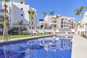 Une piscine à l’eau d’un bleu cristallin, entourée de bâtiments blancs dotés de balcons et de palmiers.
