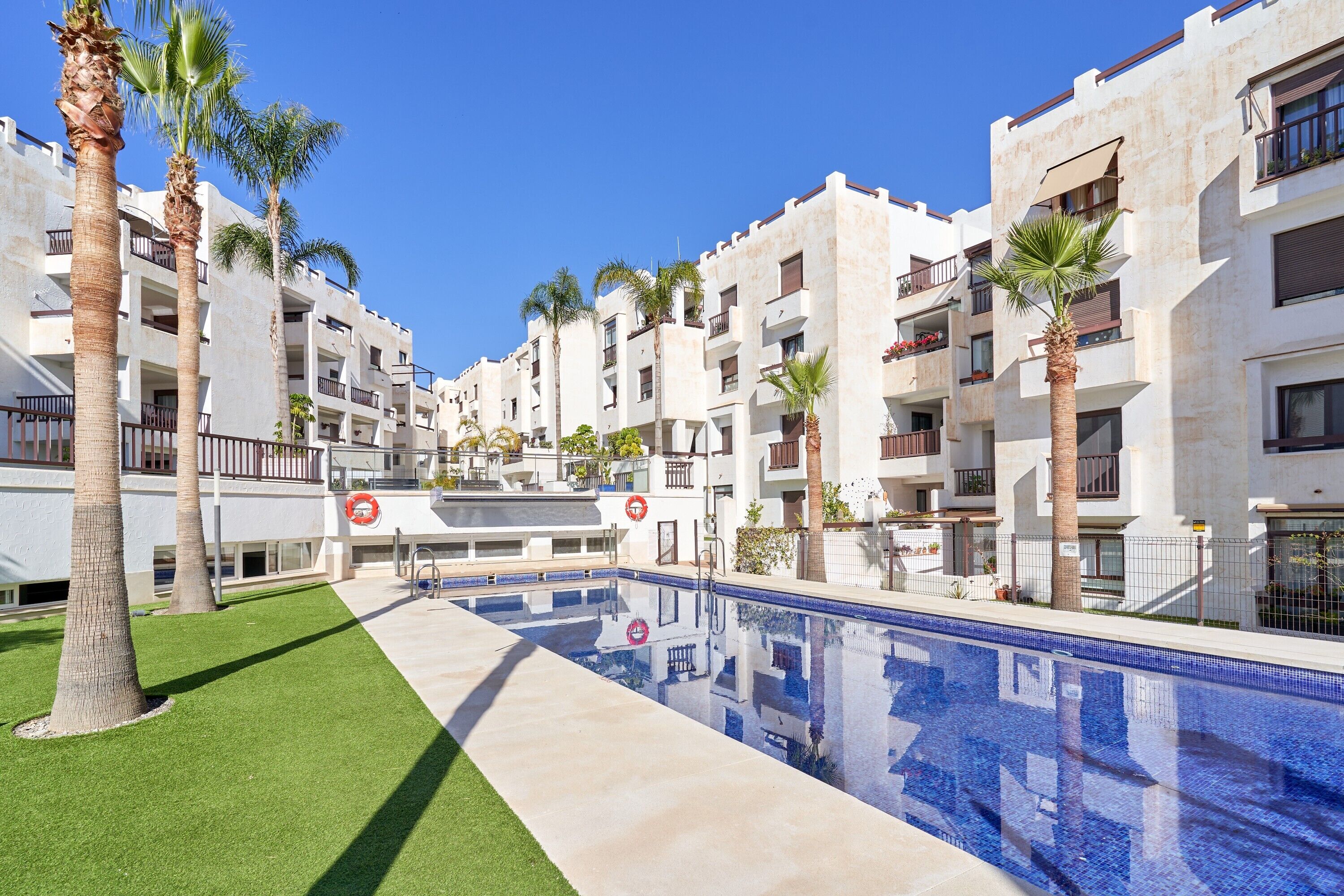 Un área de piscina con una piscina azul cristalina, rodeada de edificios blancos con balcones y palmeras.