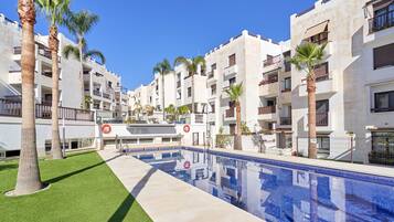 Un área de piscina con una piscina azul cristalina, rodeada de edificios blancos con balcones y palmeras.