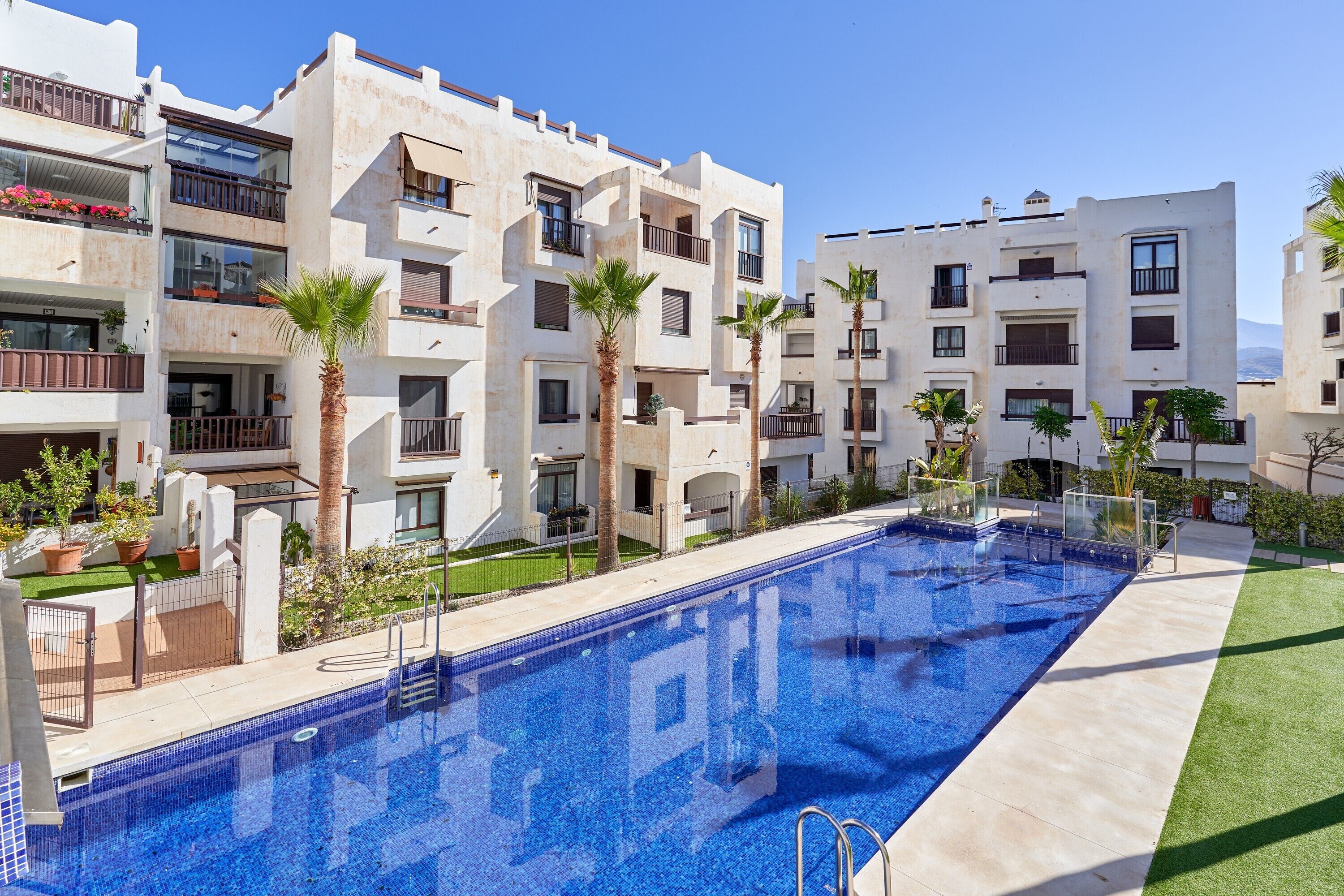 A modern hotel with a swimming pool, balconies with plants, and a clear blue sky.