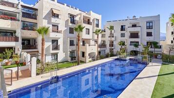 A modern hotel with a swimming pool, balconies with plants, and a clear blue sky.