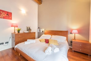 Un lit bien fait, avec du linge de lit blanc, deux lampes de chevet, une table de chevet en bois et une œuvre d’art accrochée au mur.