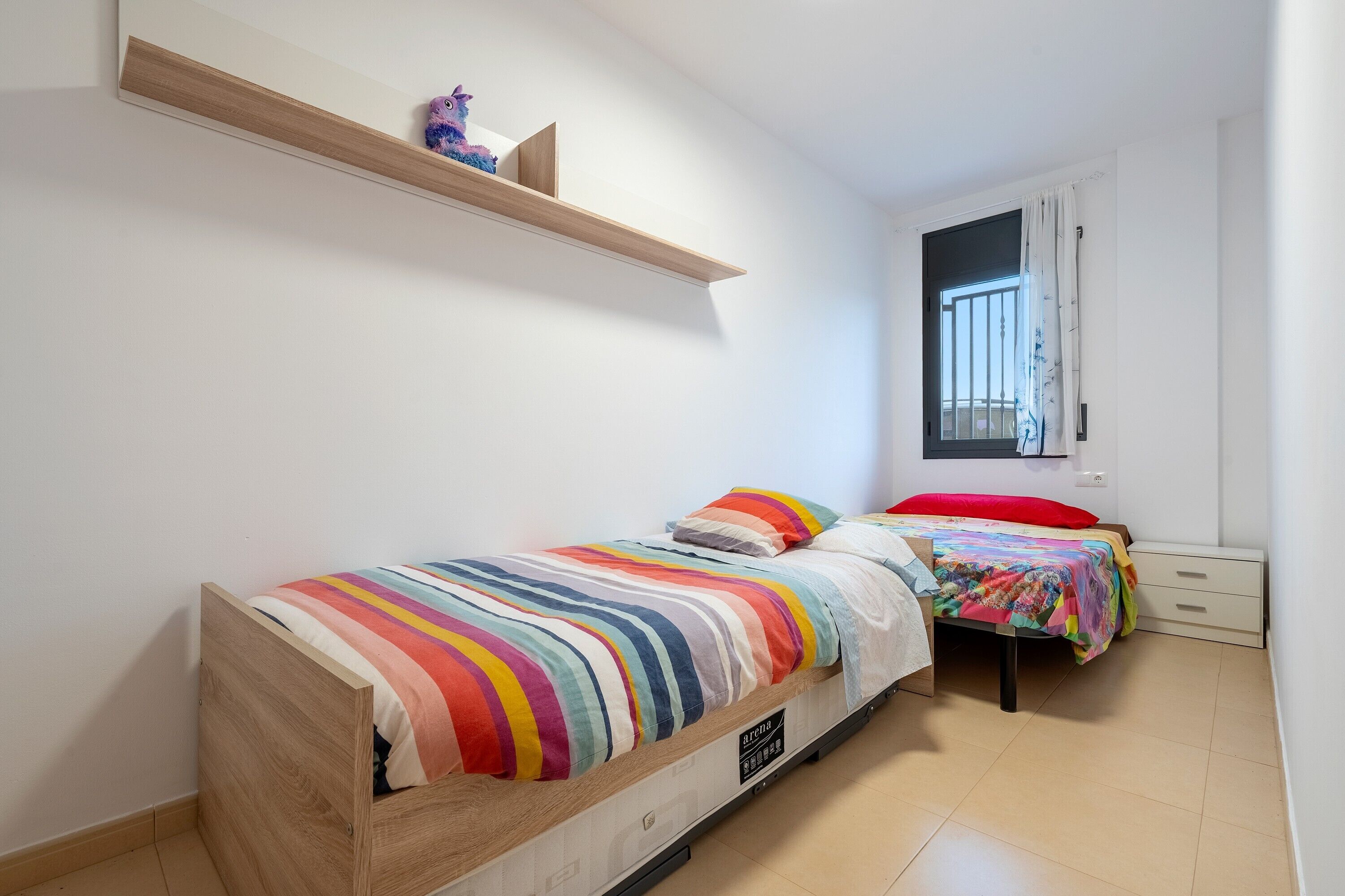 Een slaapkamer met twee bedden, een houten plank, een raam met tralies en een witte muur.