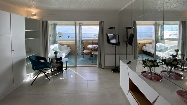 Una habitación de hotel moderna con un espejo grande, un sofá blanco, una silla azul, una mesa pequeña y vistas a la playa.