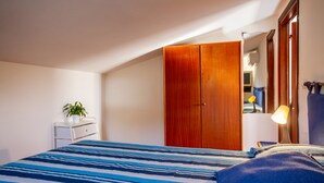 A bedroom with a bed, a wooden wardrobe, a bedside table with a lamp, and a small potted plant.