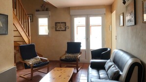 A living room with a wooden staircase, two armchairs, a sofa, a wooden coffee table, and a door with glass panels. - Holiday Home with Shared Pool, Private Terrace and Shared Garden (Saint-Trojan-les-Bains)