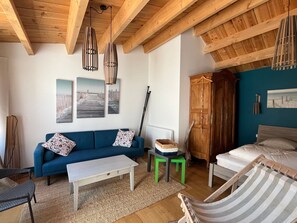 Una habitación con un sofá azul, una mesa de centro blanca, una silla a rayas, un armario de madera y una cama con ropa de cama blanca.