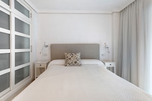 A hotel room with a large bed, two bedside tables, a nightstand, a decorative pillow, a wall-mounted lamp, and a window with curtains.