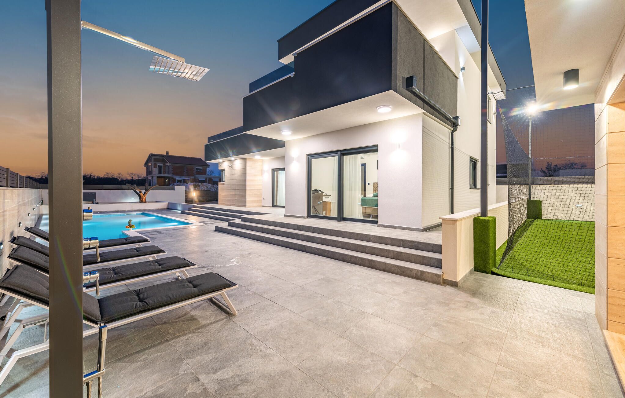 Un área de piscina moderna al aire libre con sillones de descanso, piscina y un edificio con ventanas amplias y una escalera que conduce a una zona cubierta.