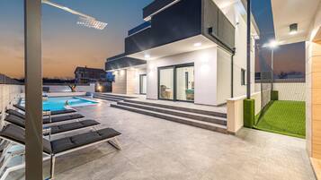 Un área de piscina moderna al aire libre con sillones de descanso, piscina y un edificio con ventanas amplias y una escalera que conduce a una zona cubierta.