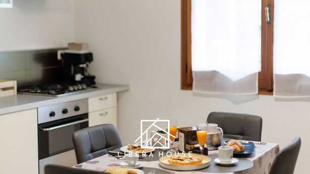 Un'area dedicata alla colazione con un tavolo apparecchiato, inclusi croissant, succo di frutta e caffè. Si intravede sullo sfondo una cucina con forno e piano cottura.