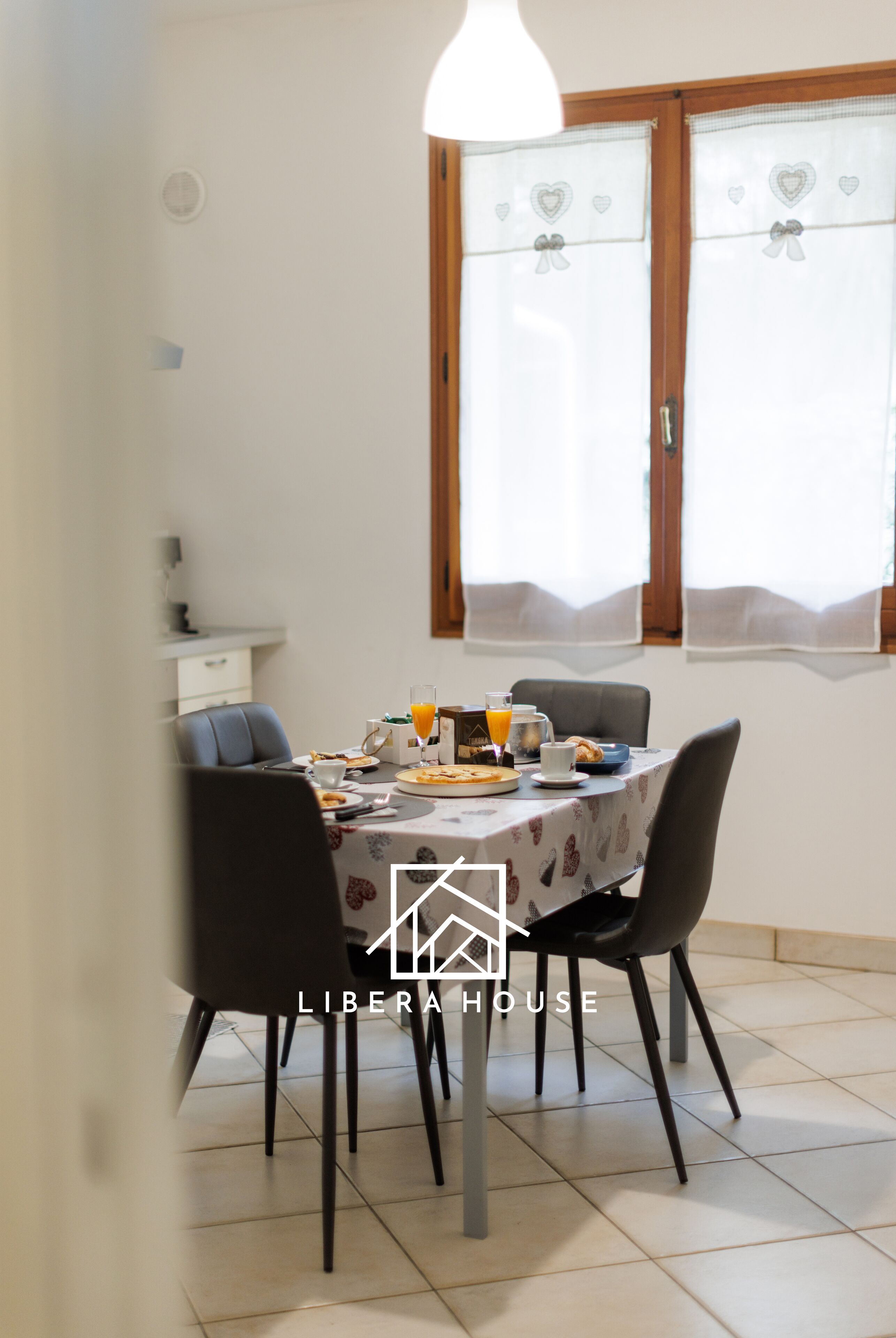 A dining area with a table set for breakfast, including plates, glasses, and a fruit bowl. There are four chairs around the table, and a window with curtains is visible in the background.