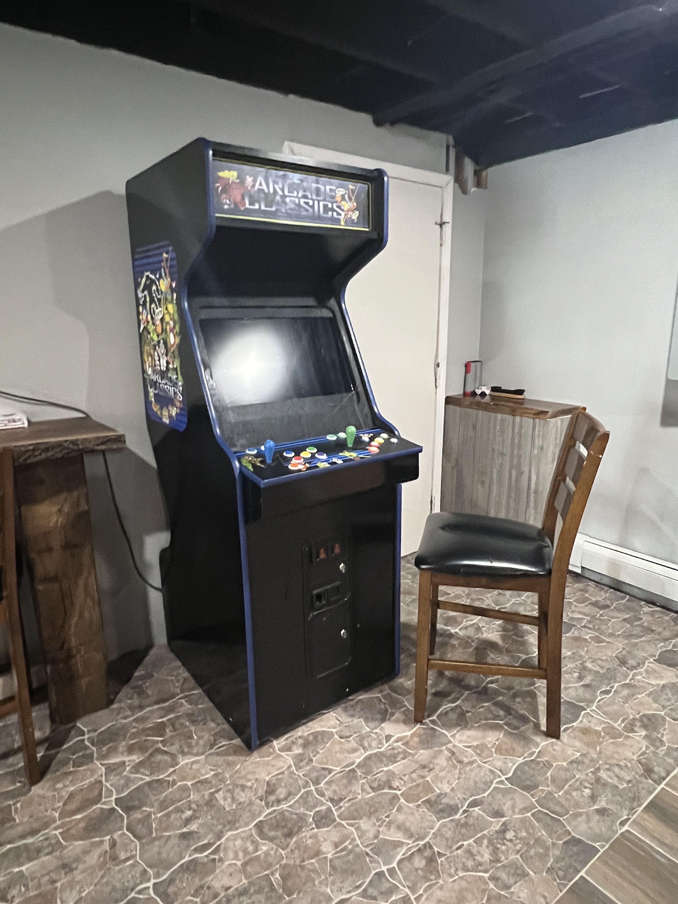Una máquina de videojuegos retro con la etiqueta "Arcade Classics", un joystick y botones, una silla de madera y un suelo con estampado de piedra.