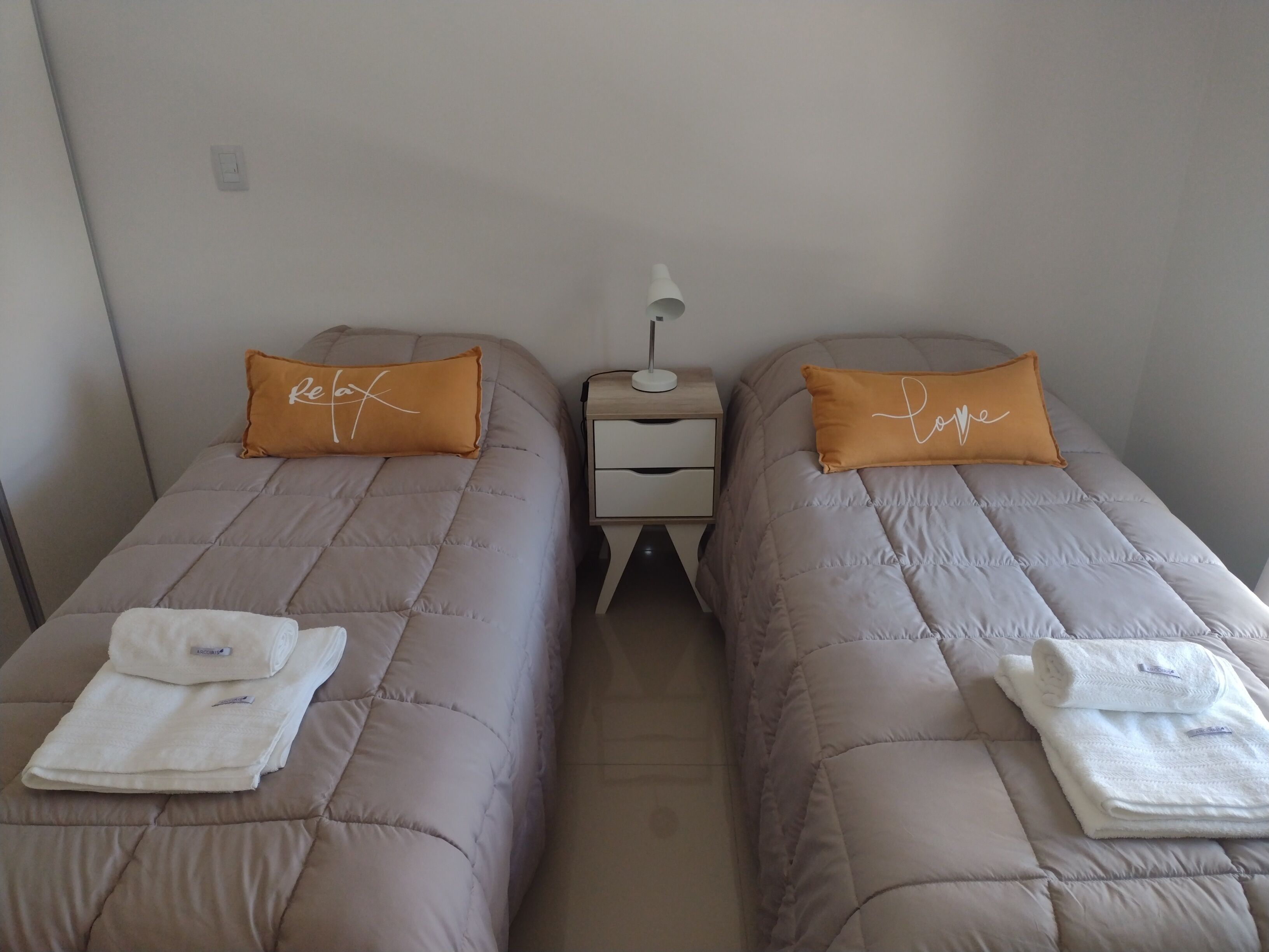 Two twin beds with beige bedding and orange pillows, each with a white towel folded on top. A small bedside table with a lamp and a drawer is between the beds.