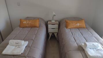 Two twin beds with beige bedding and orange pillows, each with a white towel folded on top. A small bedside table with a lamp and a drawer is between the beds.