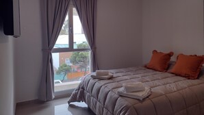 A bedroom with a bed, pillows, a window with curtains, a hanging light fixture, and a view of the outside.