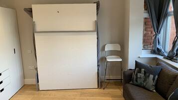 Un espacio habitable compacto con un armario blanco grande, una silla blanca pequeña, un sofá marrón con cojines y una ventana con cortinas.