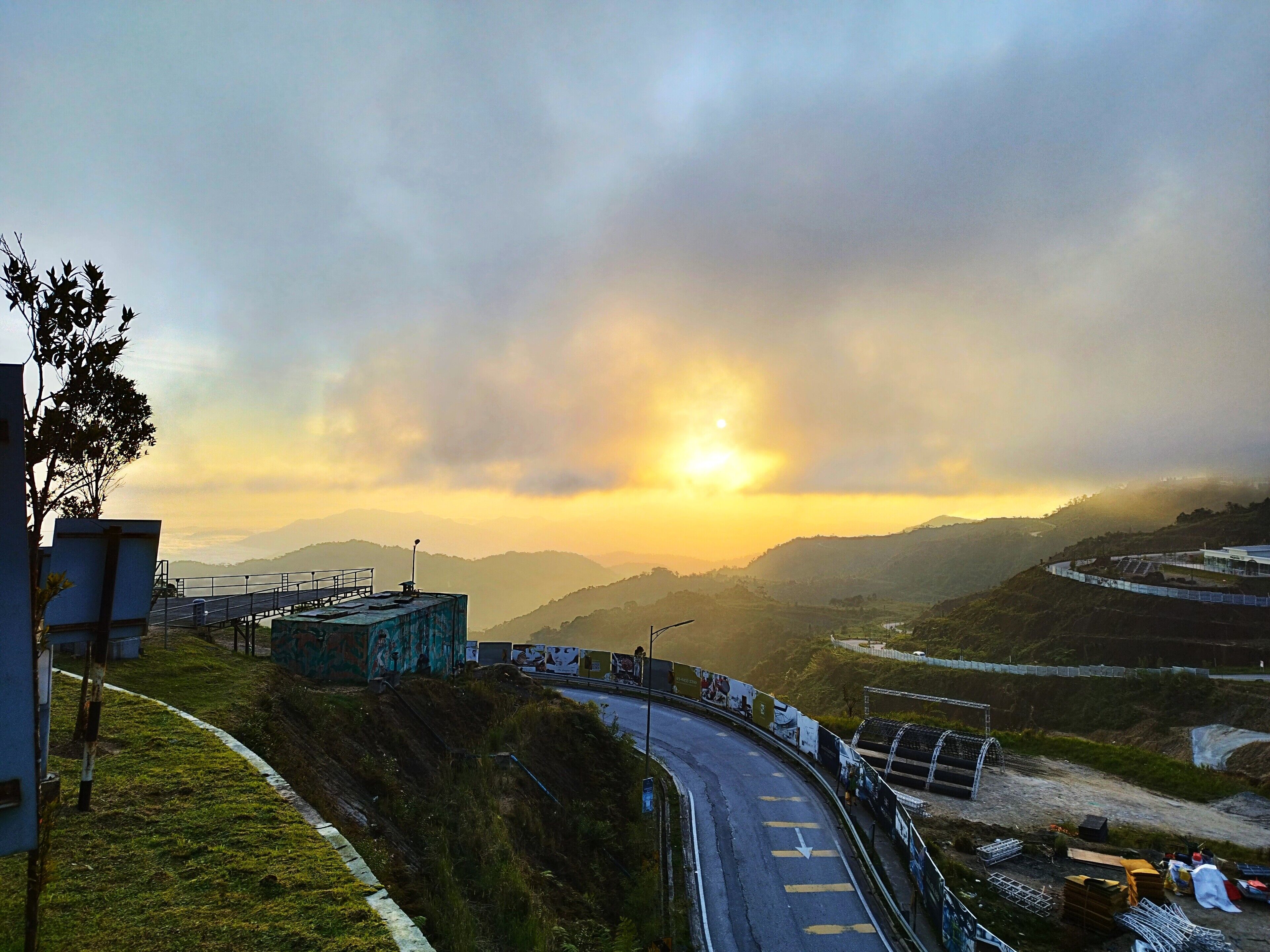 A winding road with a guardrail, surrounded by hills and a sunset in the background.