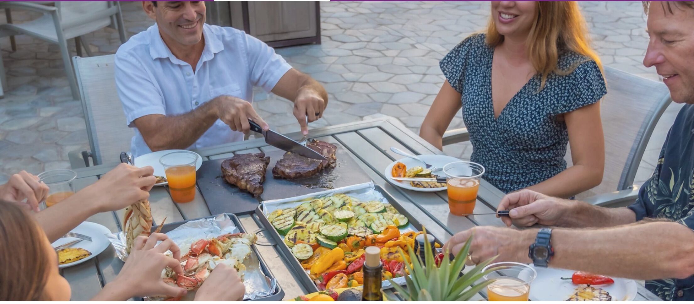 Eine Gruppe von Menschen speist draußen an einem Tisch mit gegrilltem Essen und Getränken.