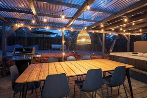 Un patio al aire libre con mesa y sillas de madera, una parrilla, un jacuzzi y luces colgantes.