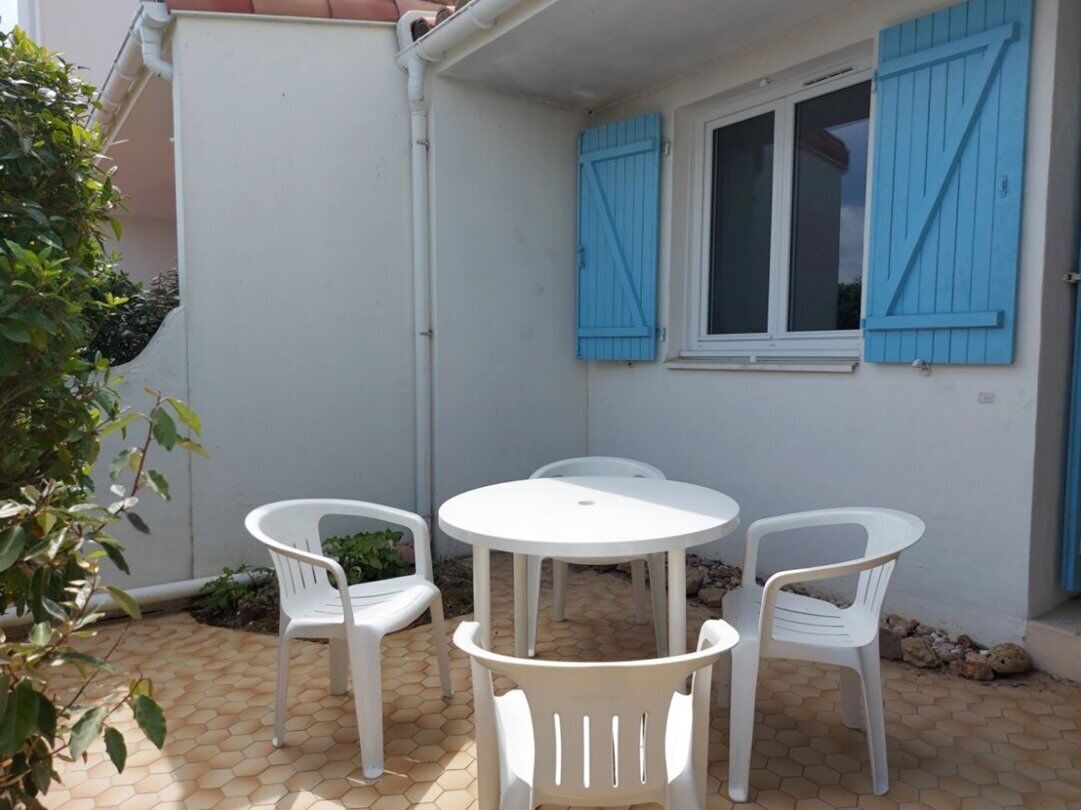 A patio with a round table and four white plastic chairs, a window with blue shutters, and a white wall.